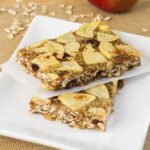 Sliced apple pie healthy oatmeal bars stacked and sitting on a white plate on a brown surface with apples and oats in the background