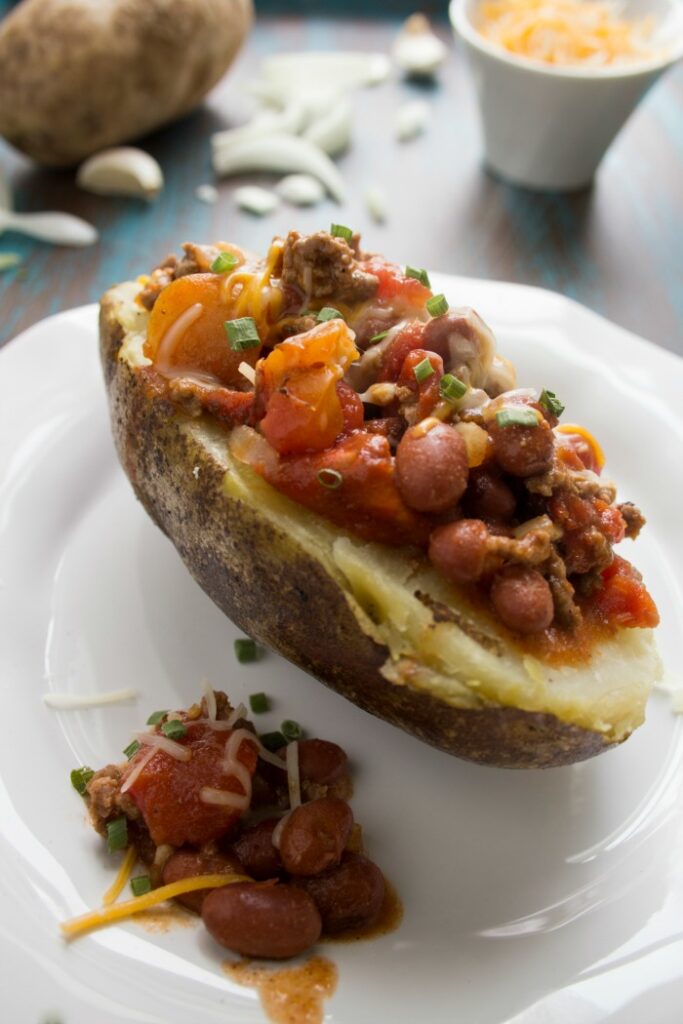 Half a baked potato on a white plate with a little chili on top and cheese in the background
