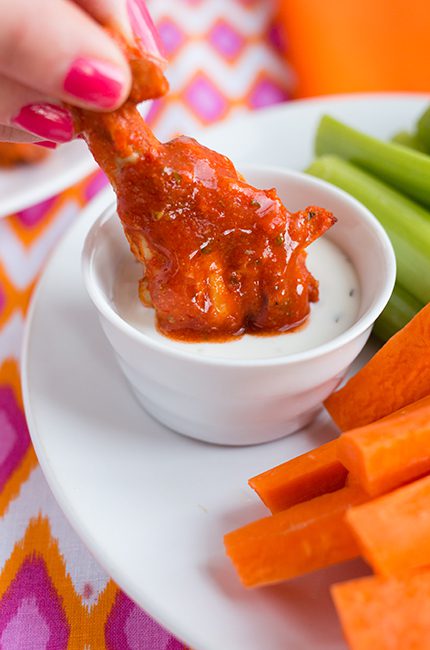 A single buffalo wing being dipped into a bowl of sauce on a plate with carrots and celery