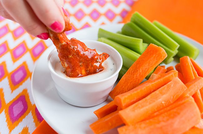 A buffalo wing being dunked into bleu cheese dressing on a plate with celery and carrots