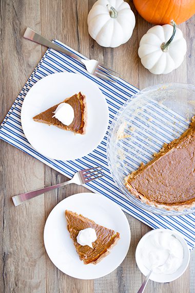 Healthy pumpkin pie slices on white plates sitting on wooden table with whipped cream on top