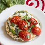 A single caprese pizza English muffin sitting on a white plate with basil in the background