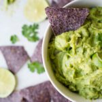 Picture of a white bowl filled with guacamole with a blue corn tortilla chip in the bowl and laying on the table aorund the bowl