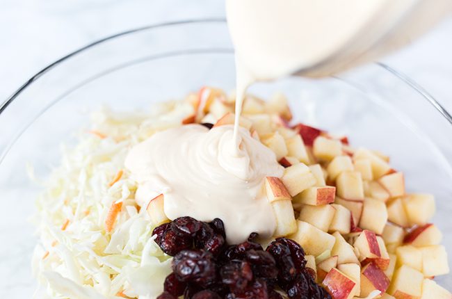 Pouring dressing over the cranberry apple coleslaw recipe in a large white glass bowl