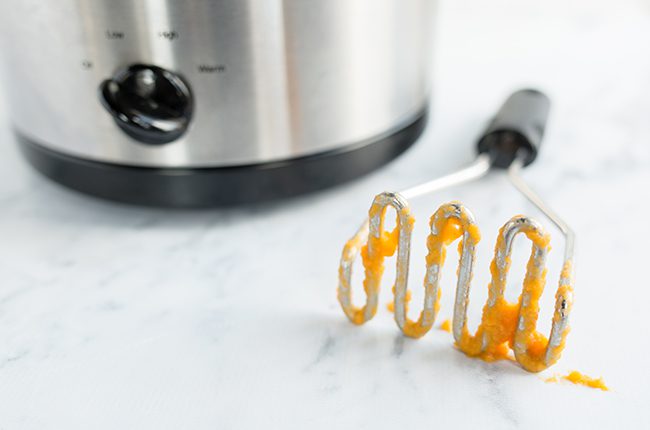 Potato masher covered in sweet potatoes sitting on counter by slow cooker