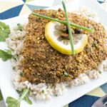 Square image of a pistachio crusted baked halibut on a white plate topped with lemon