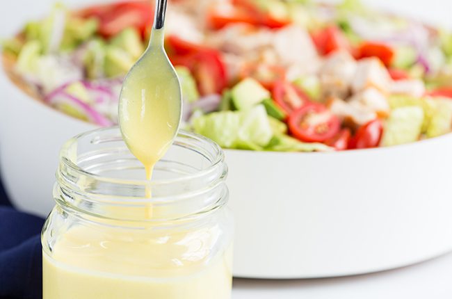 spoon drizziling honey mustard in mason jar with salad in backround