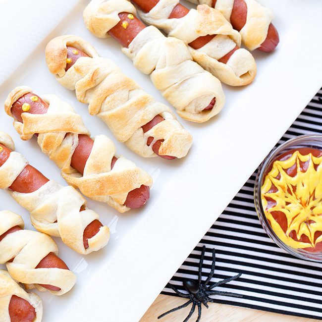 Mummy hot dogs on white serving platter with dipping sauce in background