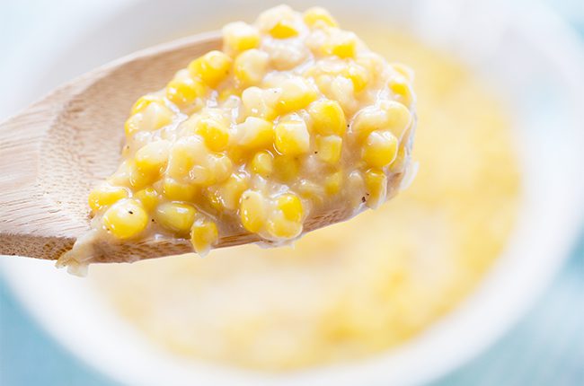 Wooden spoon with cream style corn hovering above white bowl of corn