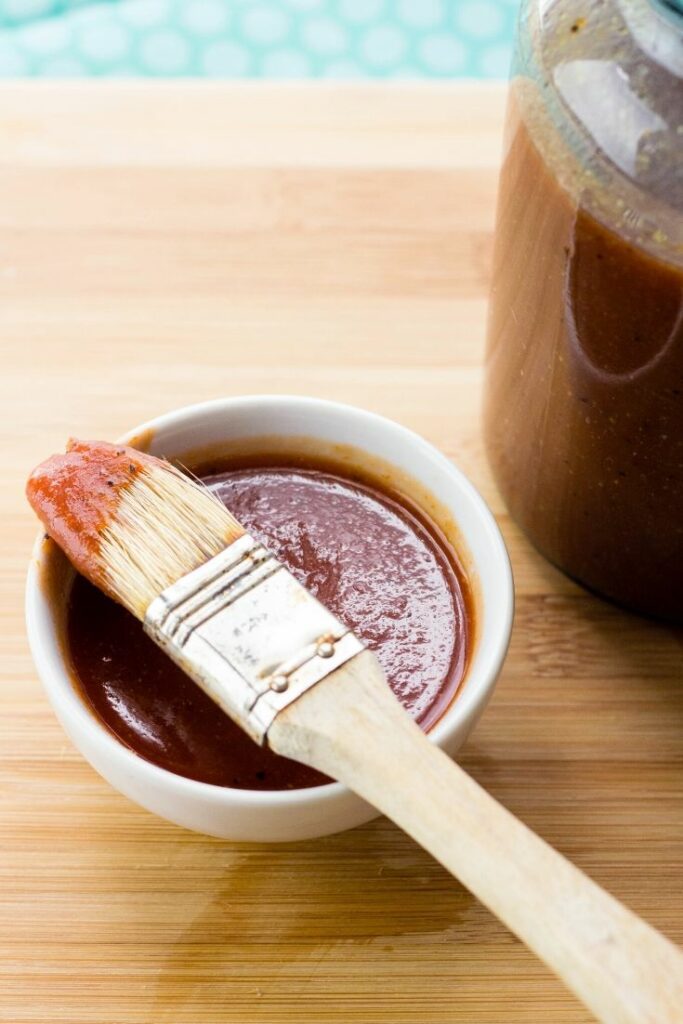 A white bowl of bbq sauce on a cutting board with basting brush