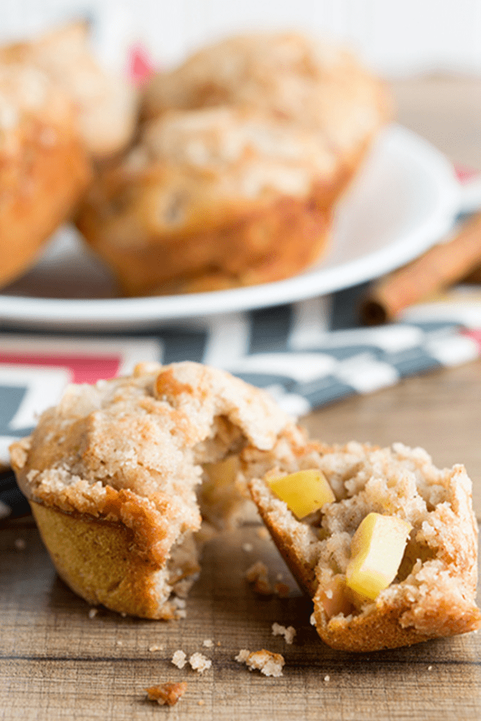 Apple cinnamon muffins split apart and sitting on wooden counter in front of plate of muffins