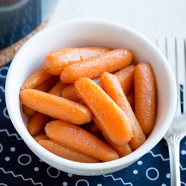 Up close overhead picture of a white bowl of candied carrots sitting on a blue and white cloth