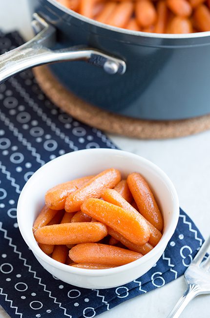A bowl of candied carrots sitting on a blue cloth in front of a black saucepan