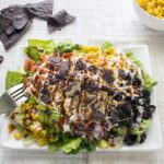 Overhead picture of a large bbq chicken salad on a white plate with ingredients on the sides