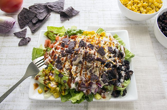 Overhead picture of a large bbq chicken salad on a white plate with ingredients on the sides