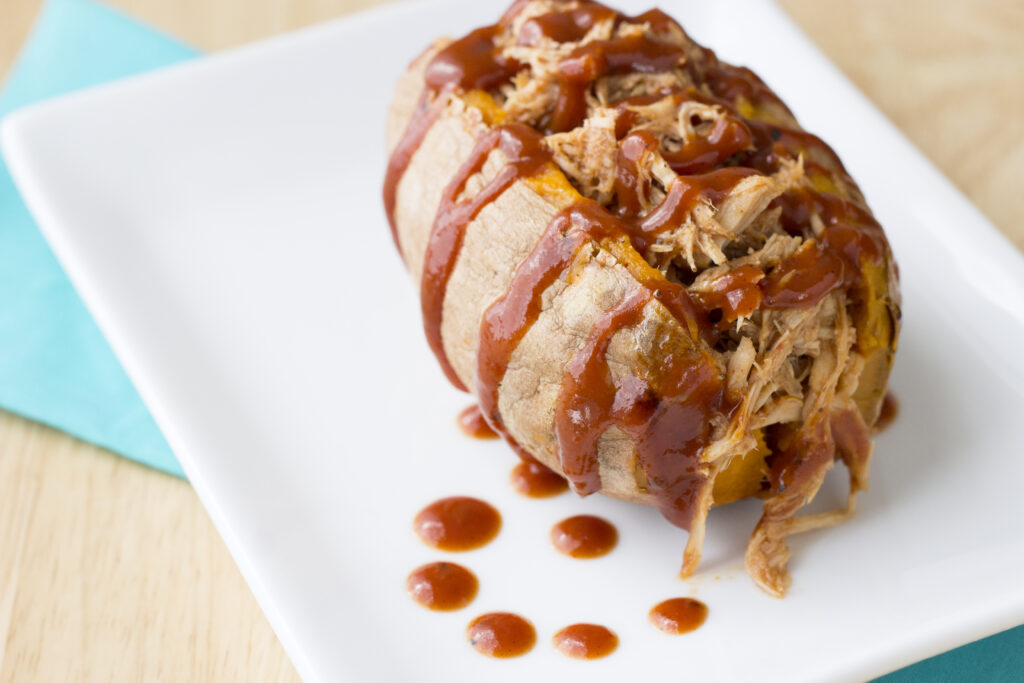 Pulled pork in baked sweet potato sitting on white plate on teal cloth napkin
