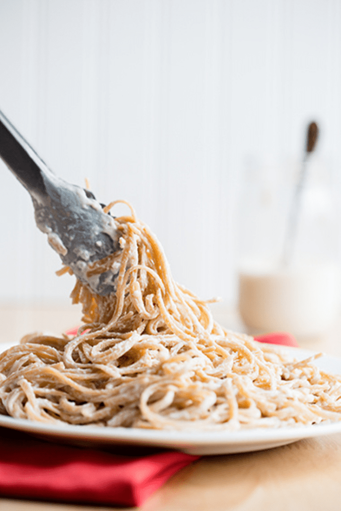 Tongs twisting whole wheat pasta coated with light alfredo sauce on a white plate