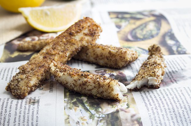 Fish sticks laying on a newspaper with lemons and tartar sauce in the background