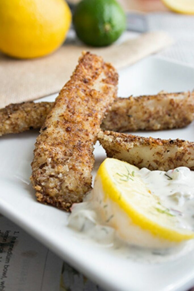 Tilapia fish sticks on a white plate with tartar sauce and lemon slices